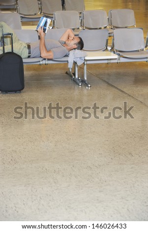 Side view of a male traveler lying on chairs and reading book in airport lobby