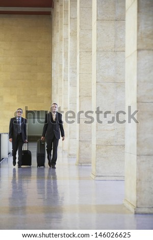 Full length of two smiling businessmen pulling luggage in airport lobby