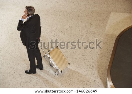 Elevated full length view of a businessman with suitcase using cellphone by luggage carousel in airport