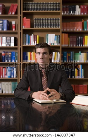 Thoughtful young man in formals with book at desk in library
