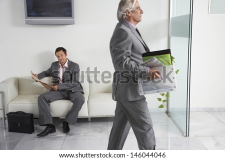 Mature businessman carrying box past a man reading newspaper in office hallway