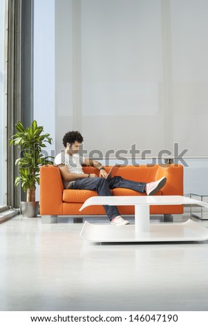 Full length of a young man using laptop on orange sofa in reception room at office