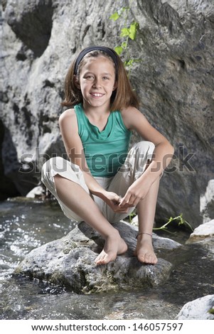 Full length portrait of a smiling little girl sitting on rock by stream