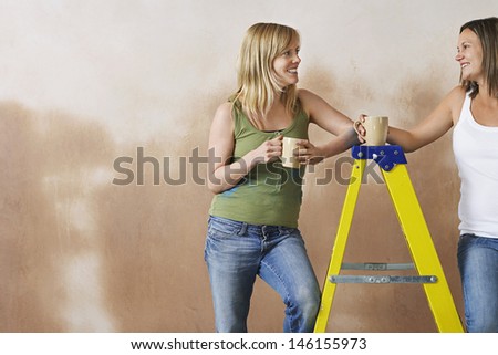 Two young women leaning on step ladder with mugs against brown wall