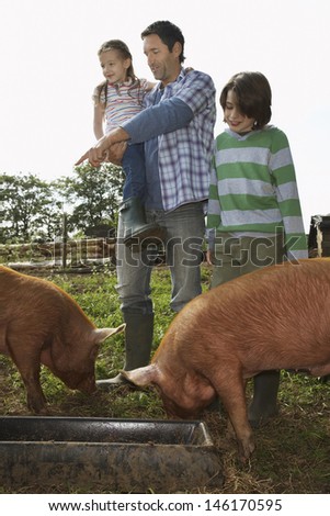 Full length of a man and children watching pigs feed in sty against clear sky