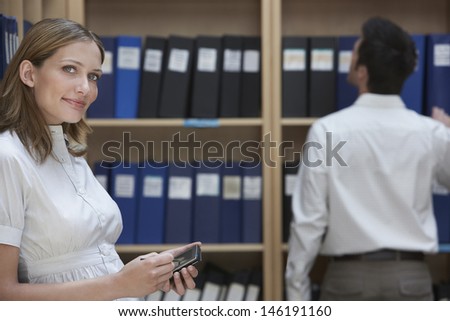 Portrait of a young office worker with palm top by male colleague in file storage room