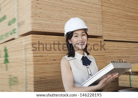 Portrait of female industrial worker holding clipboard with stacked wooden planks in background