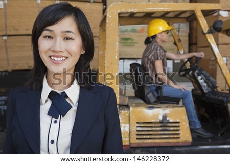 Portrait of woman smiling while female industrial worker driving forklift truck in background