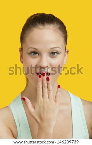 Portrait of a beautiful amazed young woman with hand over mouth over yellow background