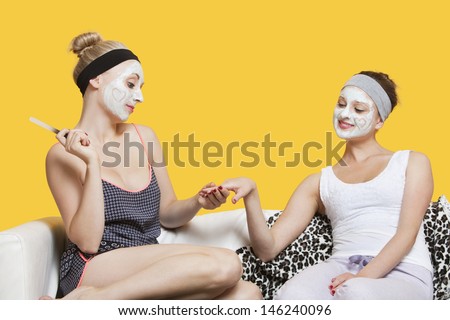 Young woman with face pack filing friend\'s nails while sitting on sofa over yellow background