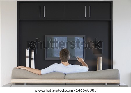 Rear View Of Young Man Using Remote Control While Sitting On Couch In Living Room