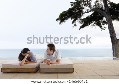Happy young couple looking at each other while relaxing on sunbeds by infinity pool