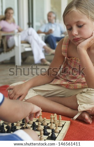 Little girl playing chess with brother at porch while parents sitting in background