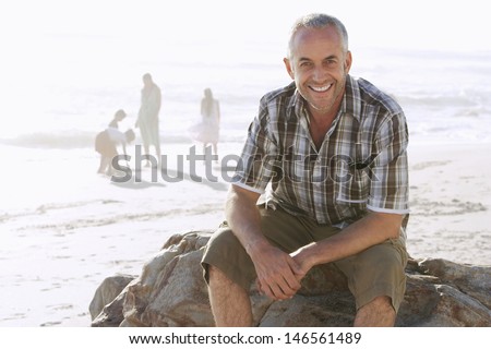 Portrait of happy middle aged man sitting on rock while family enjoying in background at beach