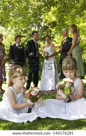 Cute little bridesmaids holding bouquets in lawn with guests and wedding couple in background