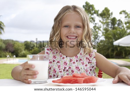 Portrait of smiling girl sitting at outdoor table with watermelon and glass of milk