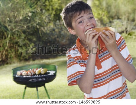 Portrait of a boy eating frankfurter with barbecue grill in the background