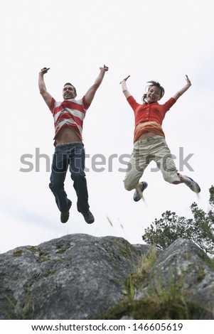 Full length low angle view of a young couple jumping with arms raised over rock
