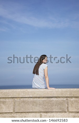 Rear view portrait of happy young woman sitting on stone ledge at beach
