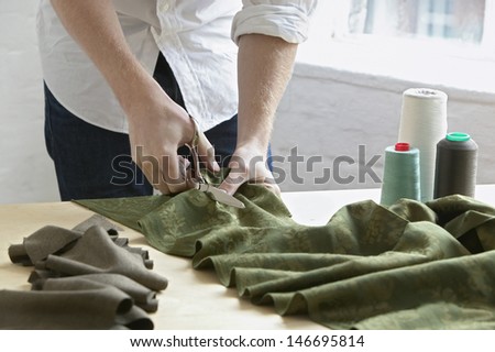 Midsection of young male tailor cutting piece of cloth at table in fashion studio