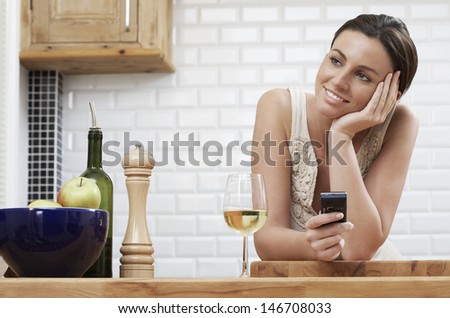 Thoughtful young woman holding cellphone while leaning on wooden counter in kitchen