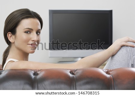 Portrait of beautiful young woman sitting on sofa with flat screen TV in background
