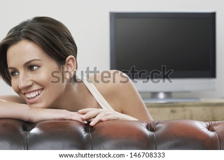 Happy young woman looking away while leaning on sofa with flat screen TV in background