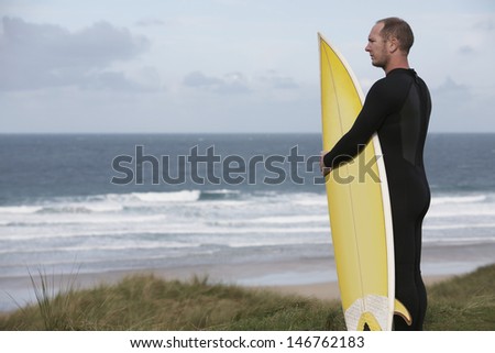 Side view of male surfer carrying surfboard on beach looking at sea