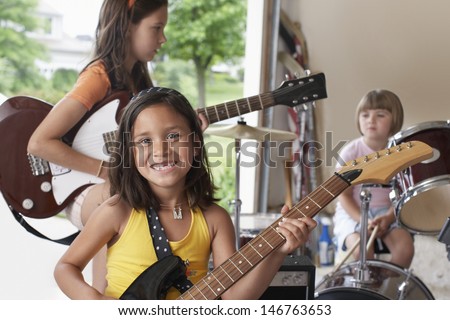 Portrait of cheerful young girl playing guitar with band in garage