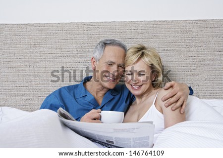 Happy middle aged couple with coffee cup reading newspaper in bed