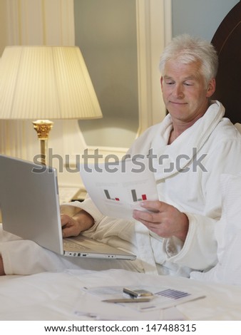 Middle aged man in bathrobe using laptop while looking at document in bed