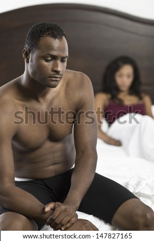 Tensed shirtless African American man sitting on bed with blurred woman in background