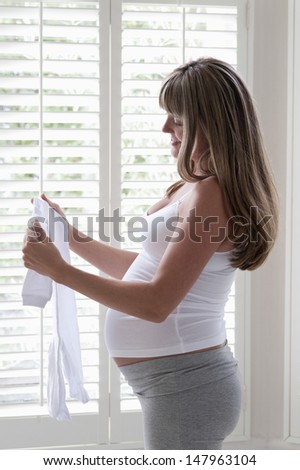 Side view of happy pregnant woman looking at baby clothing by window