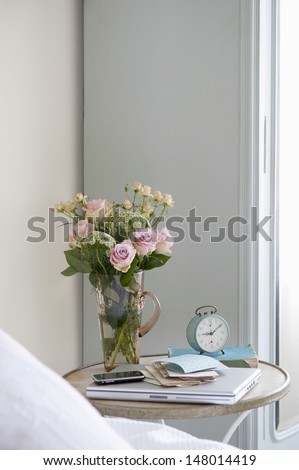 Roses In Vase On Bedside Table With Books And Alarm Clock In Bedroom