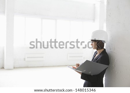 Side view of female architect leaning against wall with file folder in empty warehouse