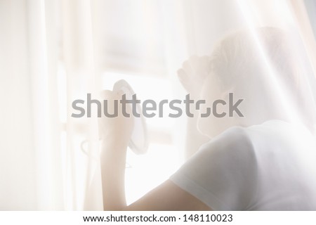 Rear view of senior woman examining herself in hand mirror behind curtains