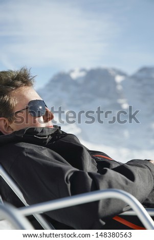 Side view of young man in winter clothes relaxing on chair against mountain