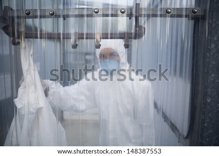 Portrait of female worker in protective mask and suit behind plastic wall at lab