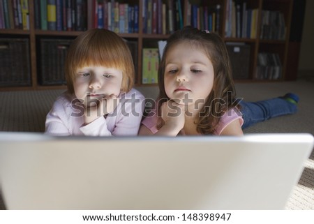 Curious young girls looking at laptop while lying on carpeted floor