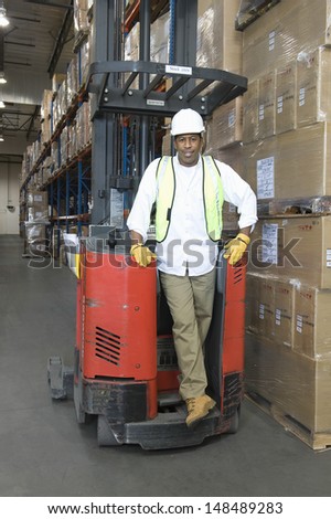 Full length portrait of a man standing with forklift truck in distribution warehouse