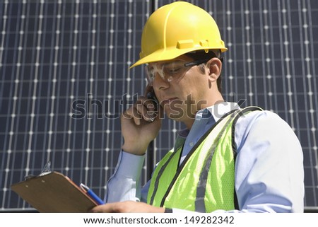 Young maintenance worker using cell phone while looking at clipboard near solar panels