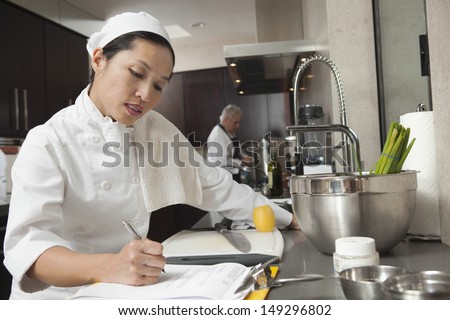 Middle aged female chef writing on clipboard with colleague working in background