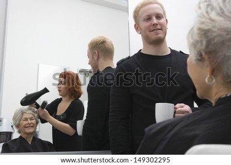 Stylist blow drying senior woman\'s hair while man holding coffee cup in salon