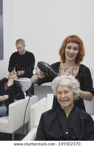 Happy female stylist blow drying senior woman\'s hair in salon