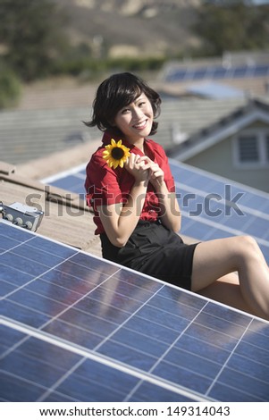 Beautiful young woman holding sunflower while sitting by solar panel on rooftop