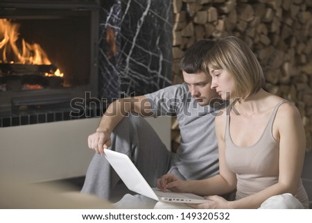 Young couple using laptop while sitting by fireplace at house