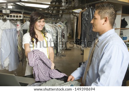 Young female owner showing dry cleaned clothes to customer at counter in laundry
