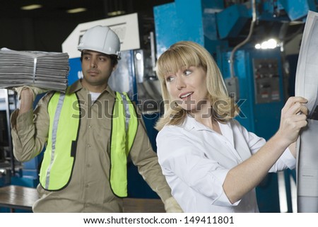 Woman checking newspaper with colleague in the background in factory