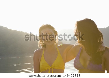 Closeup portrait of two cheerful young women in bikinis with arms around against the lake