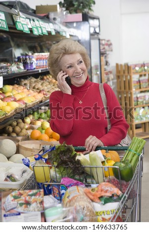 Smiling mature woman using cellphone while doing grocery shopping in supermarket